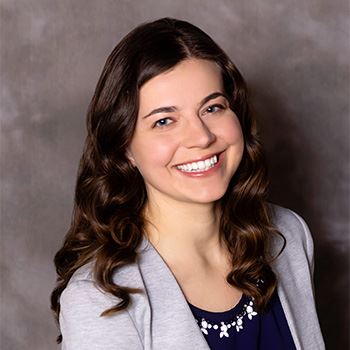 Casey Casella wears a navy blouse with a gray blazer. Her brown hair reaches just past her shoulders